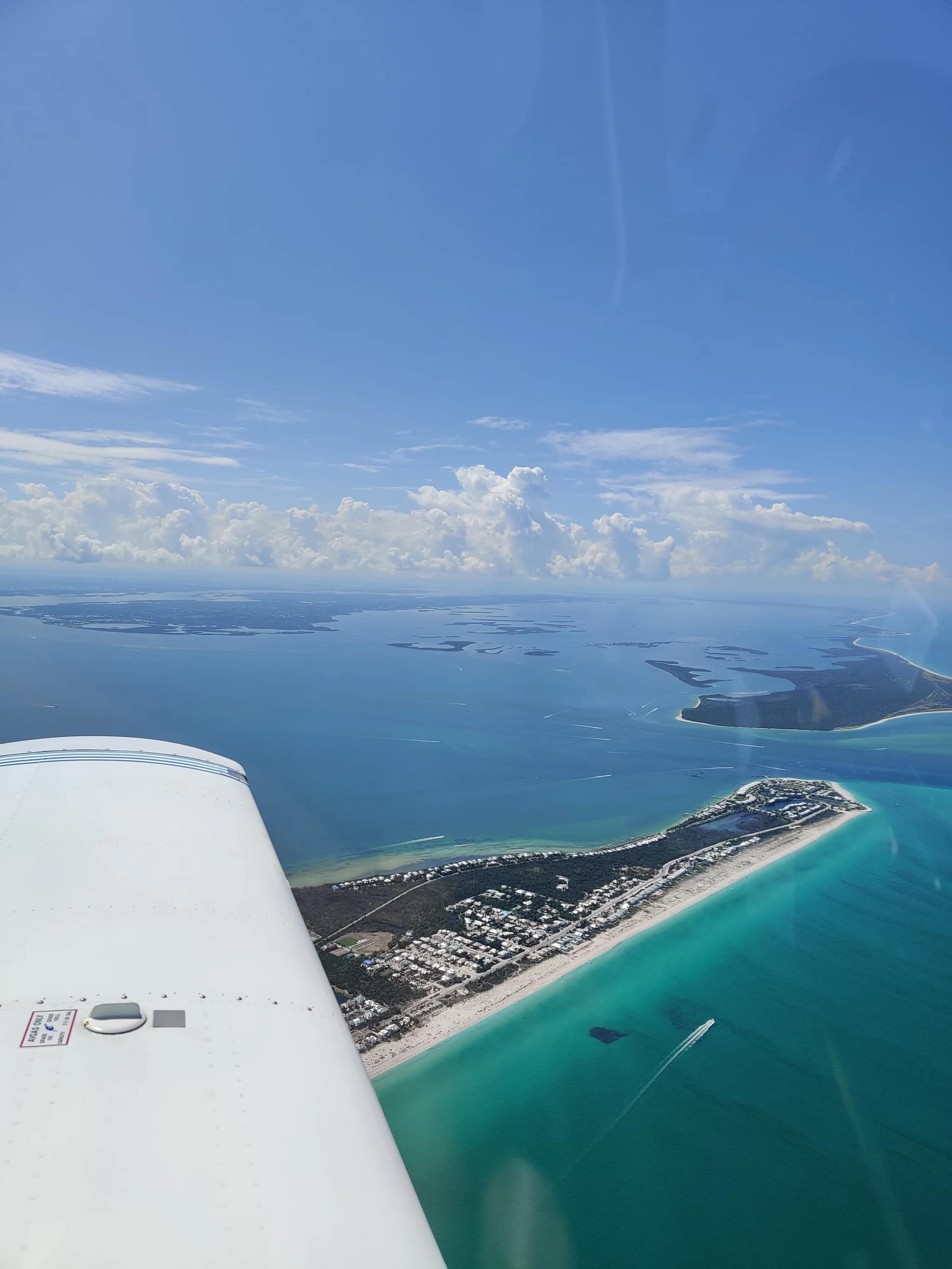 View over the wing of a low-wing trainer: turquoise Gulf water, a white-sand barrier island, and the Southwest Florida coastline