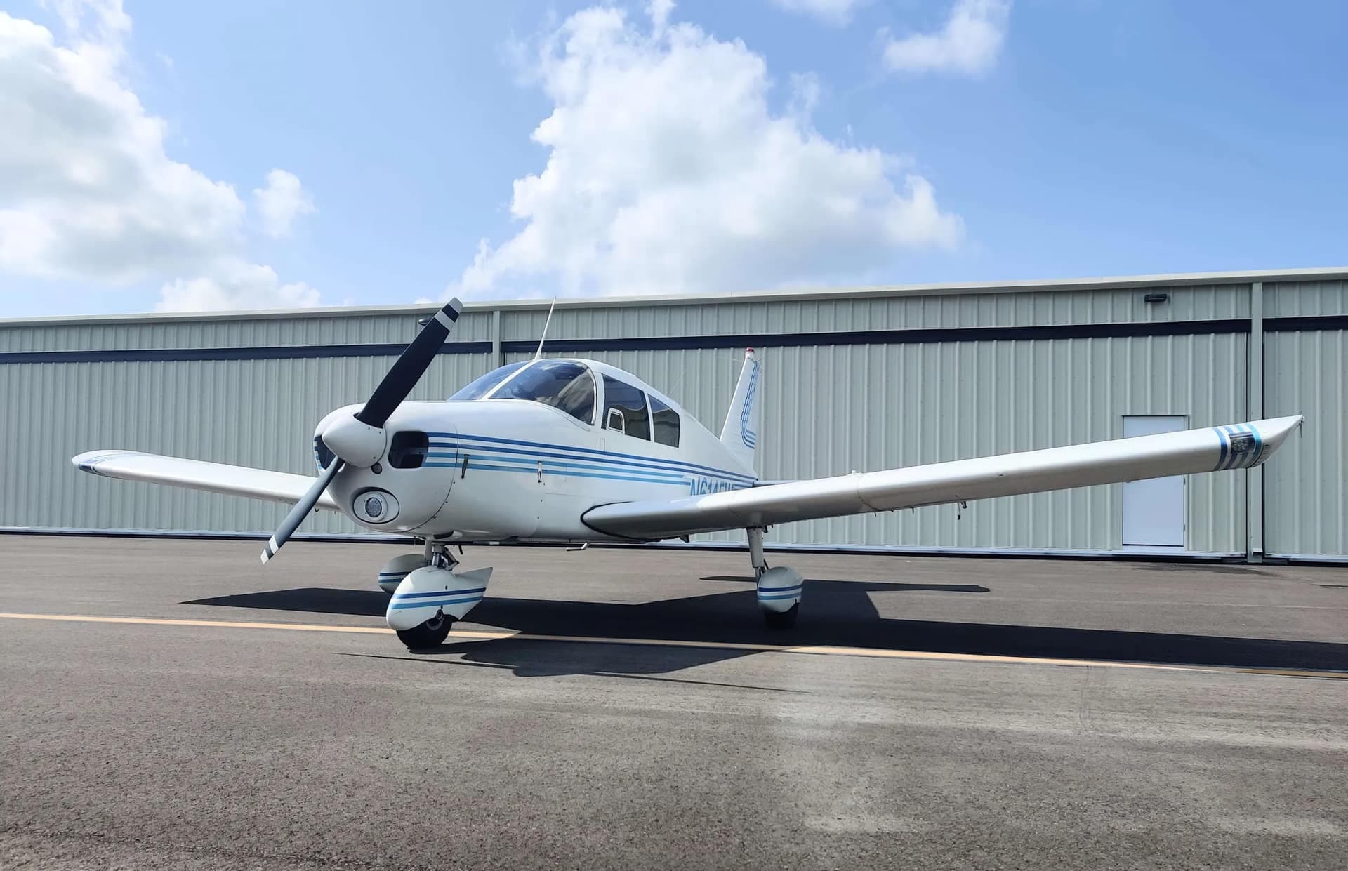 White-and-blue Piper Cherokee 140 parked on the ramp in front of a hangar under a blue Florida sky